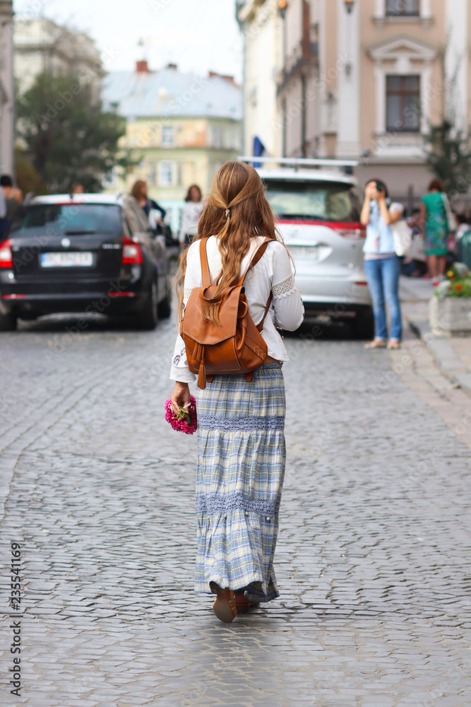 A girl with long, loose hair walks in beautiful hippie style clothes along the ancient streets of a medieval city. Young tourist in fashionable stylish clothes handmade with a knapsack and a flowers