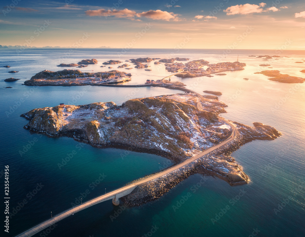 Aerial view of bridge over the sea and snowy mountains in Lofoten Islands, Norway. Henningsvaer at sunset in winter. landscape with blue water, sky with clouds, rocks, buildings, road. Top view