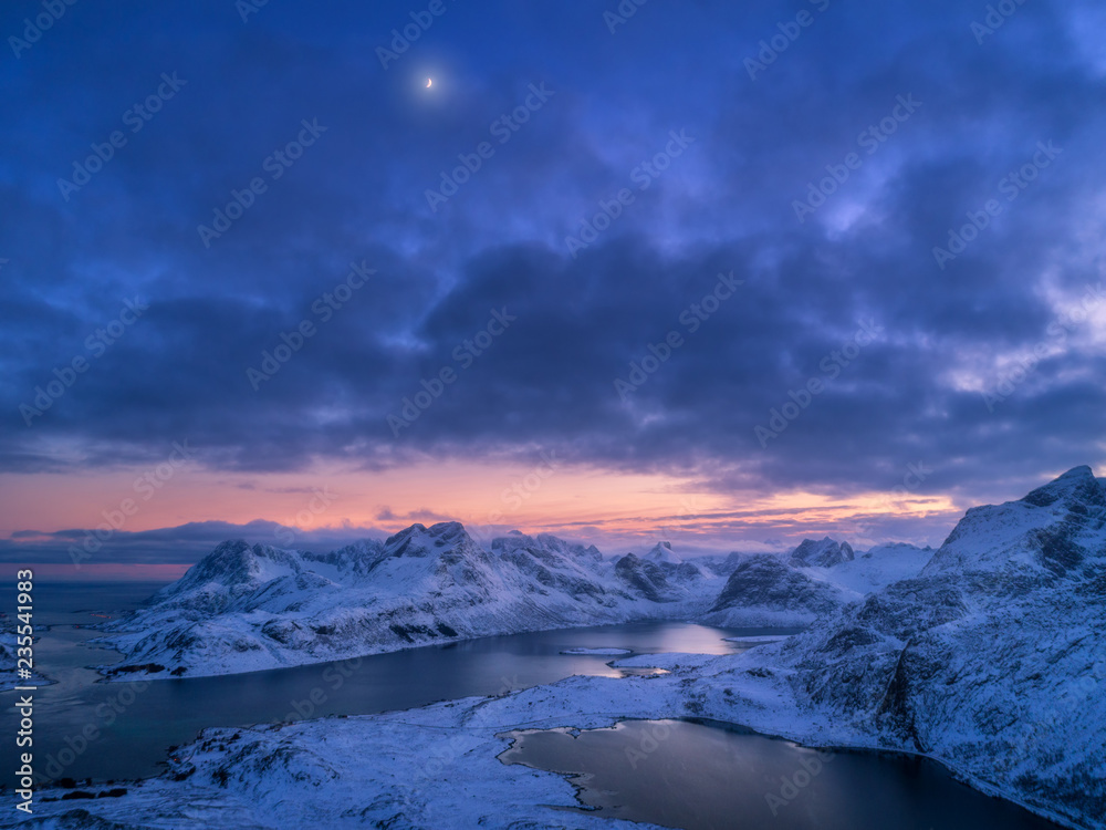 Aerial view of snowy mountains, sea, colorful cloudy sky at night in Lofoten islands, Norway. Winter landscape with snow covered rocks and seacoast and sunset sky. Top view of Norwegian Fjords at dusk