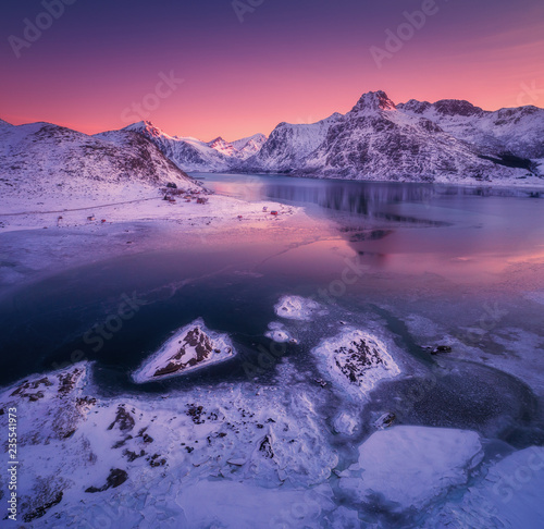 Aerial view of snowy mountains  blue sea with frosty coast  reflection in water and purple sky at colorful sunset in Lofoten islands  Norway. Winter landscape with snow covered rocks  fjord with ice