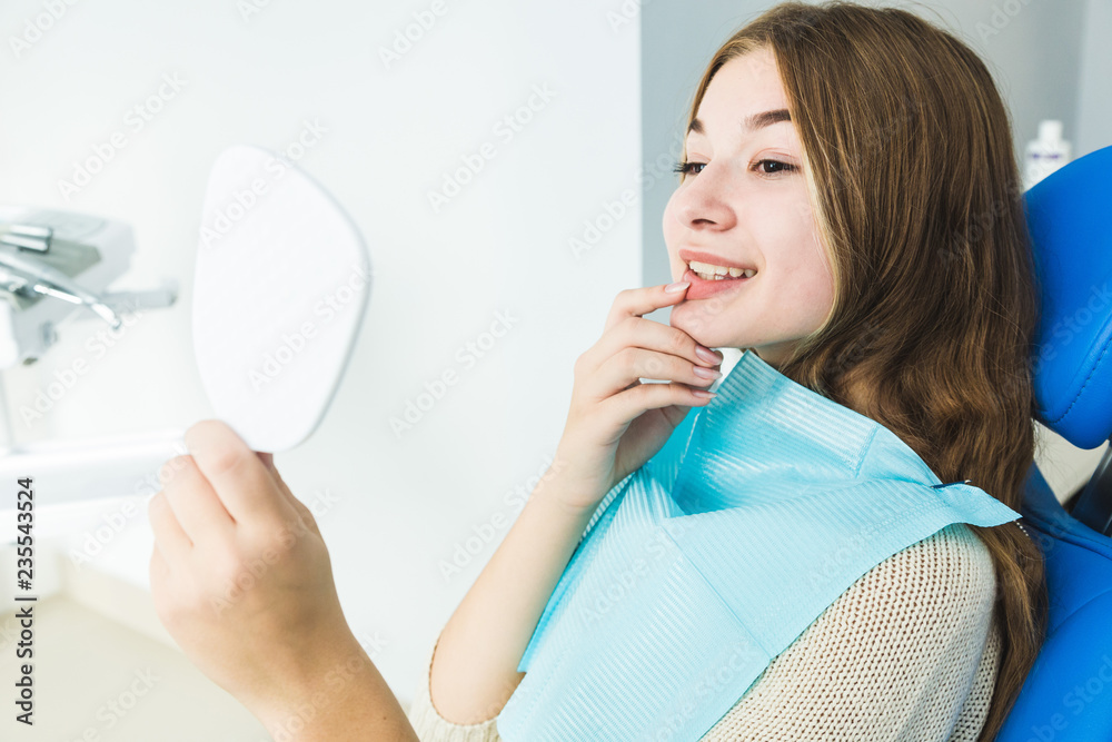 Dental clinic. Reception, examination of the patient. Teeth care. Young girl smiling, looking in the mirror after a dental checkup at her dentist Dental clinic. Reception, examination of the patient. Teeth care. Young girl smiling, looking in the mirror after a dental checkup at her dentist