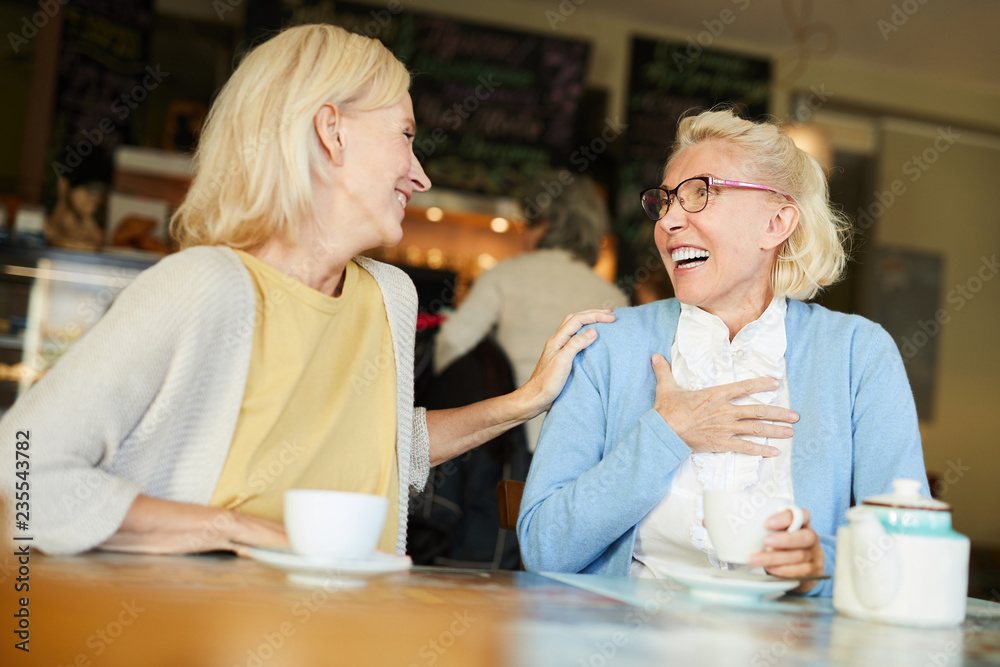 Two female friends laughing during their talk by cup of tea while relaxing in cafe at leisure