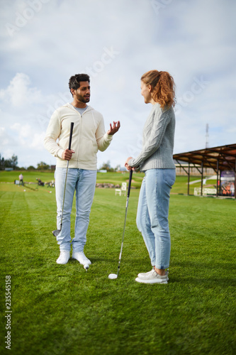 Young man and woman in casualwear discussing golf play stuff on large green field