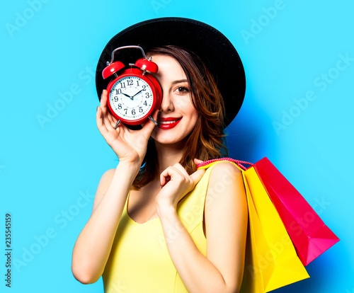 Portrait of young smiling red-haired white european woman in hat with alarm clock and shopping bags on blue background