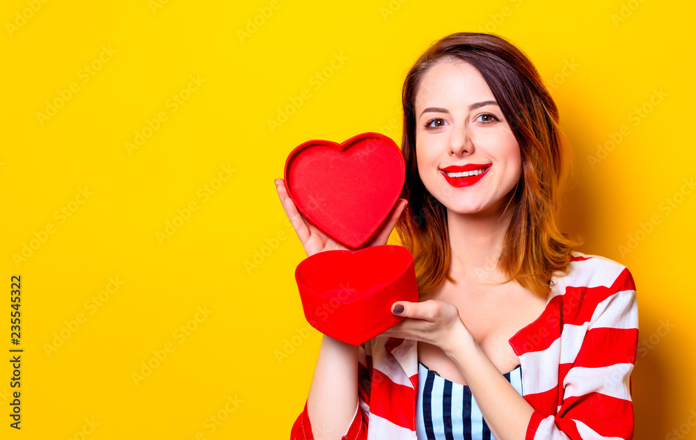 Portrait of beautiful young smiling red-haired white european woman in red striped shirt with heart shape box on yellow background Portrait of beautiful young smiling red-haired white european woman in red striped shirt with heart shape box on yellow background