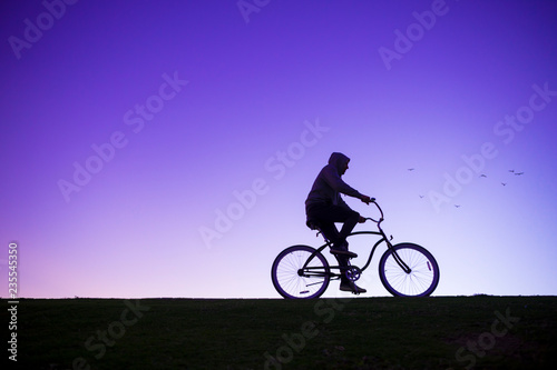 Silhouette of man in hoodie riding a beach cruiser bicycle against a glowing purple sky