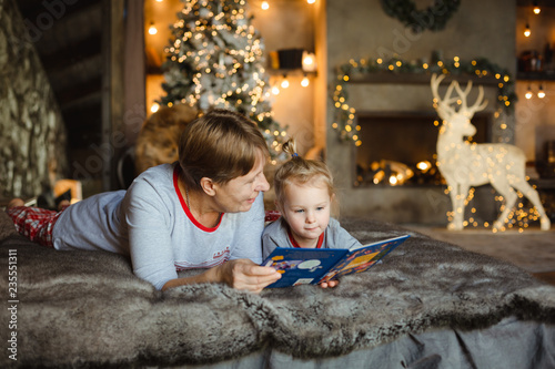 Grandmother and granddaughter in Christmas pajamas reading a book, lying on the bed in the Chalet, decorated for Christmas - Christmas tree and garlands around the fireplace. Family Christmas concept.