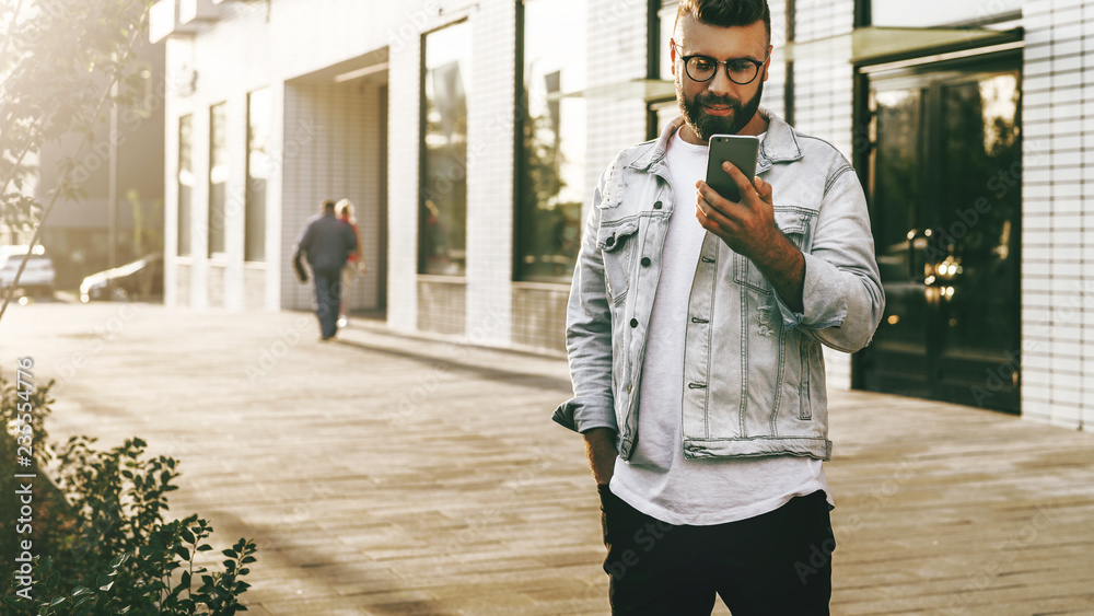 Fototapeta premium Handsome smiling hipster businessman with beard, in denim jacket, trendy glasses walks around city and using smartphone.
