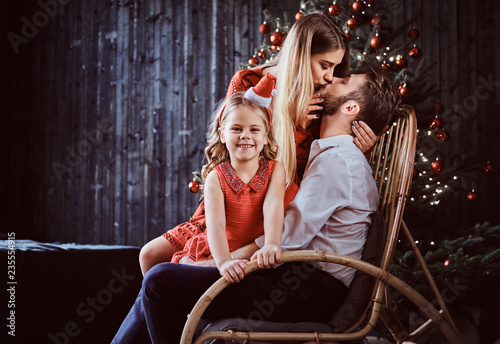 A happy little girl looking at a camera while her parents kiss on a rocking chair near a Christmas tree at home.