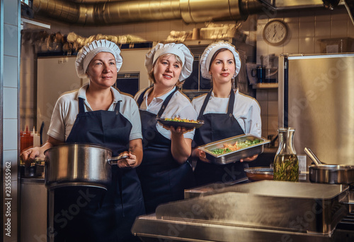 Cooking team wearing a uniform holding containers with chopped vegetables, posing for a camera at restaurant's kitchen.