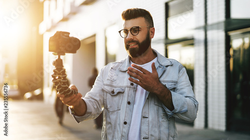 Bearded male hipster blogger in trendy glasses standing on city street, holds camera on tripod and shoots video blog. photo