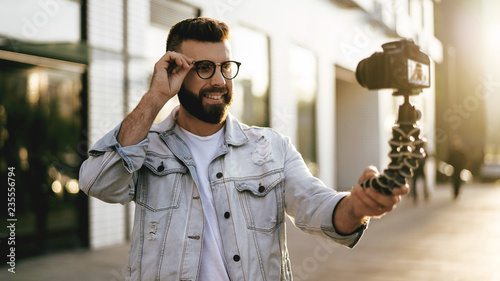 Bearded male hipster blogger in trendy glasses standing on city street, holds camera on tripod and shoots video blog. photo