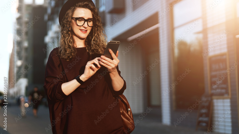 Obraz premium Young curly hipster woman in trendy glasses and hat walks on city street and uses smartphone. Social media,lifestyle.