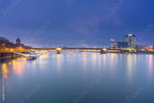 Blick auf den wunderschönen Rhein, Schiffe die sich im Wasser spiegeln, die Deutzer Brücke und die beleuchtete Skyline in Köln bei Nacht,