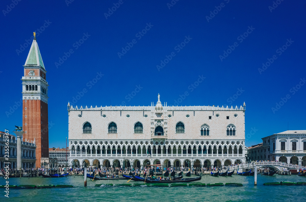 Facade of Ducal Palace, St. Mark's Bell Tower over water and gondolas, under blue sky, in Venice, Italy