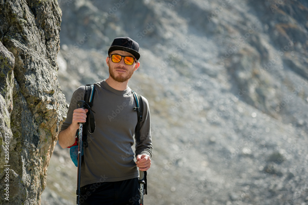 Porter of a stylish hipster traveler with a beard and a backpack in sunglasses and a cap with trekking poles stands on a rock against the backdrop of a mountain circus Porter of a stylish hipster traveler with a beard and a backpack in sunglasses and a cap with trekking poles stands on a rock against the backdrop of a mountain circus