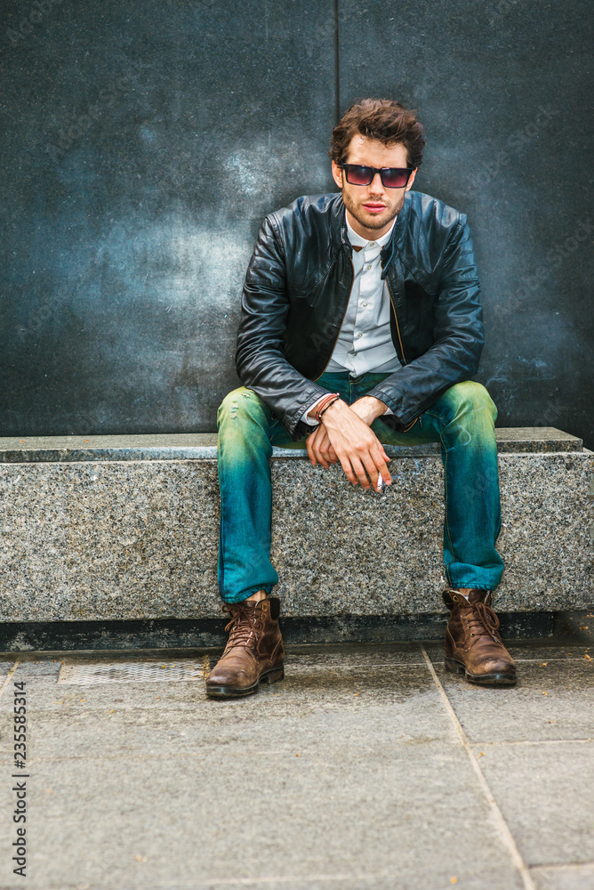 Wearing black leather jacket, blue jeans, brown boot shoes, sunglasses, a  young guy with beard sitting on marble bench in corner, smoking cigarette  during working break, trying mind calming down Stock Photo