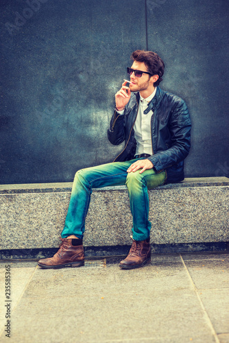 Wearing black leather jacket, blue jeans, brown boot shoes, sunglasses, a young guy with beard sitting on marble bench in corner, smoking cigarette during working break, trying mind calming down