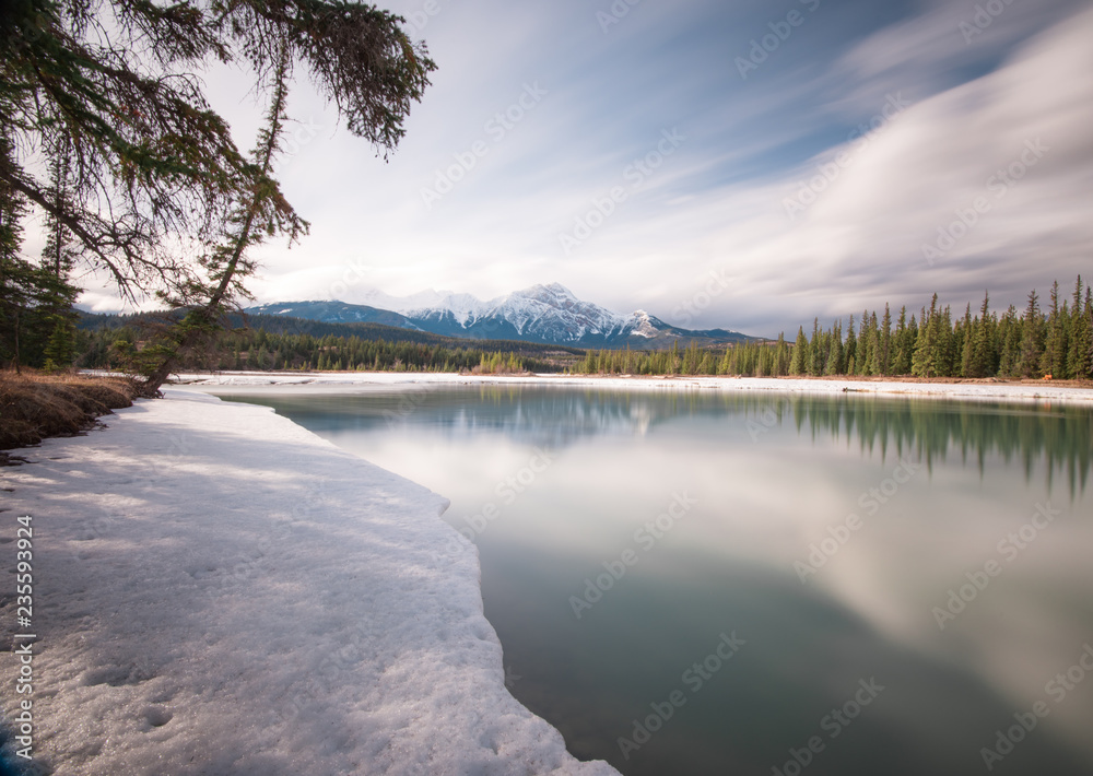 Trees overhang the snow covered edged of a thawing river in early spring in an alpine setting Trees overhang the snow covered edged of a thawing river in early spring in an alpine setting