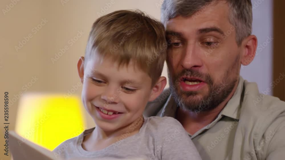 Close-up of cheerful little boy smiling and bonding with dad reading a fairytale together in the evening Close-up of cheerful little boy smiling and bonding with dad reading a fairytale together in the evening