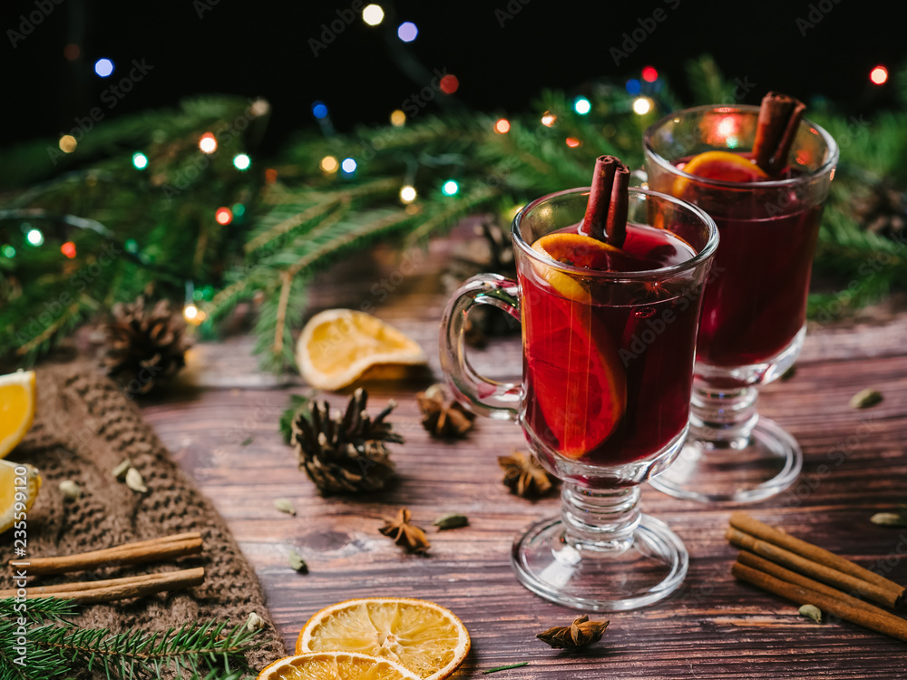 mulled wine in glass glasses with orange, cinnamon, cardamom, a anise stars on the table decorated with a Christmas tree and a garland. Traditional hot new year's eve alcoholic drink mulled wine in glass glasses with orange, cinnamon, cardamom, a anise stars on the table decorated with a Christmas tree and a garland. Traditional hot new year's eve alcoholic drink