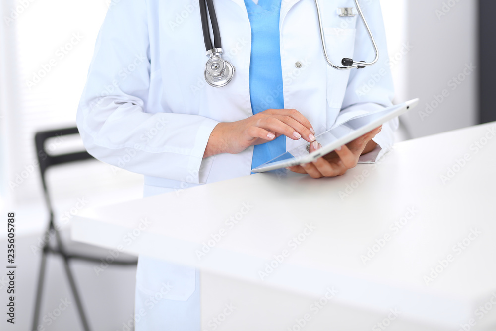 Close-up of female physician using digital tablet while standing near reception desk at clinic or emergency hospital. Unknown doctor woman at work. Medicine and healthcare concept Close-up of female physician using digital tablet while standing near reception desk at clinic or emergency hospital. Unknown doctor woman at work. Medicine and healthcare concept