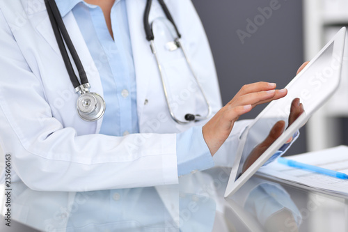 Close-up of female physician hands using digital tablet  while sitting at glass desk at hospital office. Medicine and healthcare concept