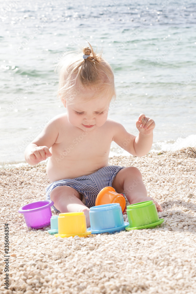 Cute baby playing with plastic toys on the pebble beach near the sea Cute baby playing with plastic toys on the pebble beach near the sea