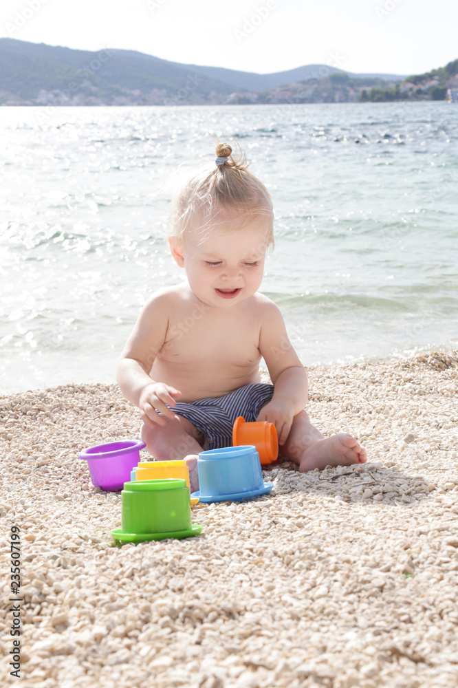 Cute baby playing with plastic toys on the pebble beach near the sea Cute baby playing with plastic toys on the pebble beach near the sea