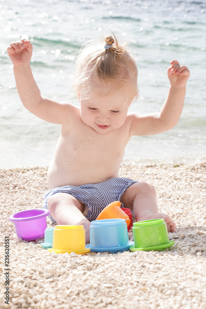 Cute baby playing with plastic toys on the pebble beach near the sea Cute baby playing with plastic toys on the pebble beach near the sea