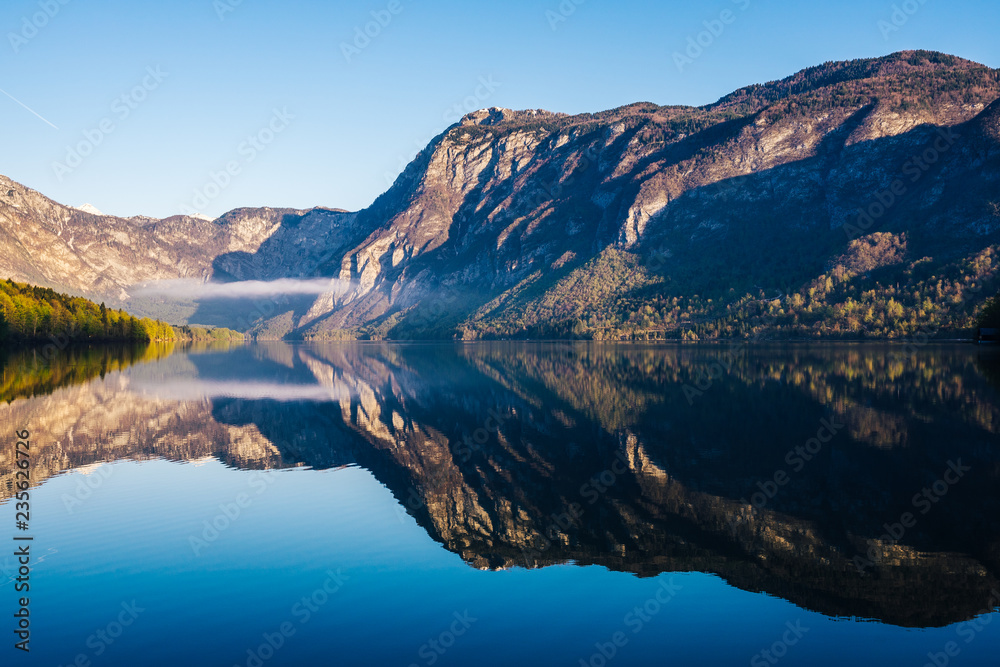 Obraz premium Sunrise view of alpine lake Bohinj with blue sky, sunlight and mist over water. Circles in water. Famous lake of Bohinj in Triglav National Park, Julian alps, Slovenia.