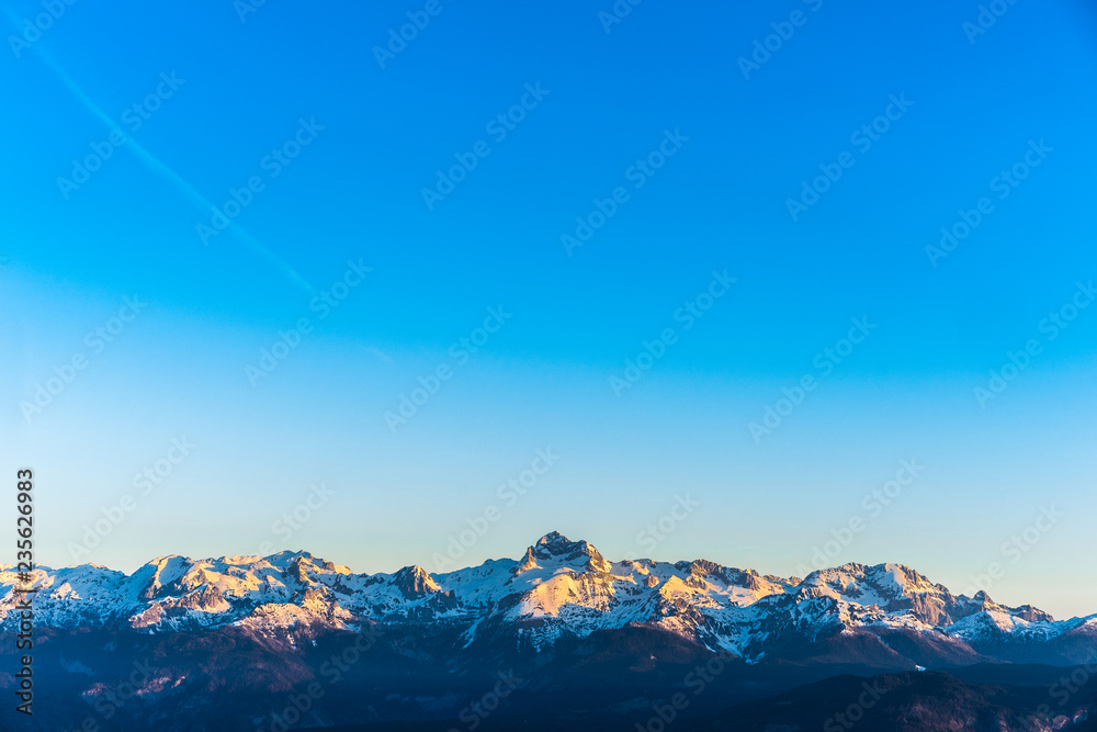 Sunrise over the Julian alps and the highest mountain of Slovenia Triglav. Blue sky and alpine landscape on horizont with sun shining on snow covered summit of Mount Triglav. Wallpaper or background.