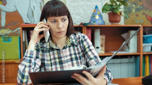 A young female teacher dressed in a checkered shirt sits at a table in the classroom, she checks the presence of characters in the folder for the game and talking on the phone.