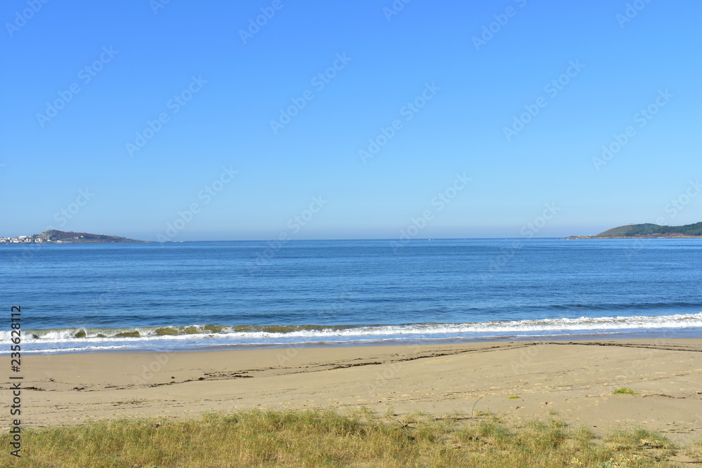 Beach with golden sand and vegetation in sand dunes. Blue sea with small waves and foam. Sunny day, Galicia, Spain.
