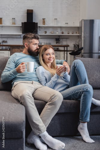 Cheerful couple sitting on sofa in casual clothing with cups