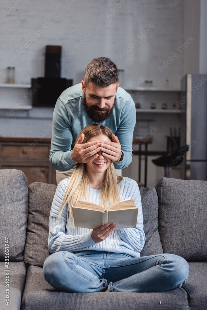 Cheerful husband closing eyes of happy wife with book