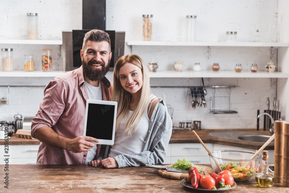happy couple holding digital tablet with blank screen at wooden kitchen table happy couple holding digital tablet with blank screen at wooden kitchen table