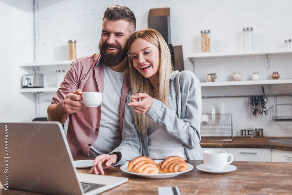 smiling woman pointing at laptop screen while man drinking coffee smiling woman pointing at laptop screen while man drinking coffee