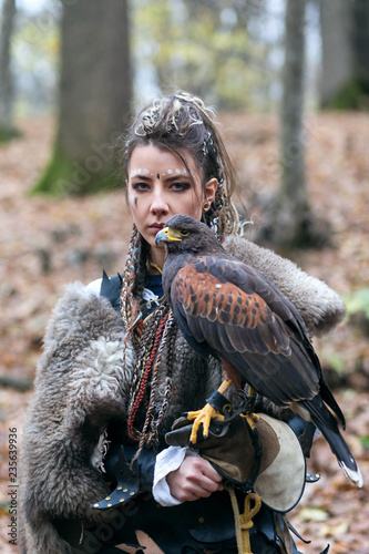 Foto Outdoor portrait of Viking warrior woman in woods wearing fur collar, with braided hair and specific makeup with face covered in blood holding hawk in hand