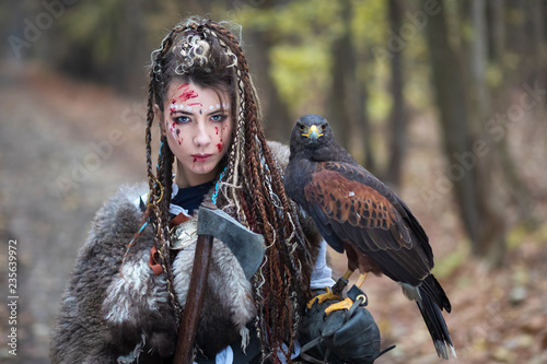 Schilderij op canvas Beautiful Viking warrior woman in woods wearing fur collar, braided hair and specific makeup with face covered in blood holding hawk in hand