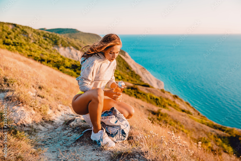 Woman with bottle of water in nature at warm sunset. Attractive girl hiker in mountains and ocean at background