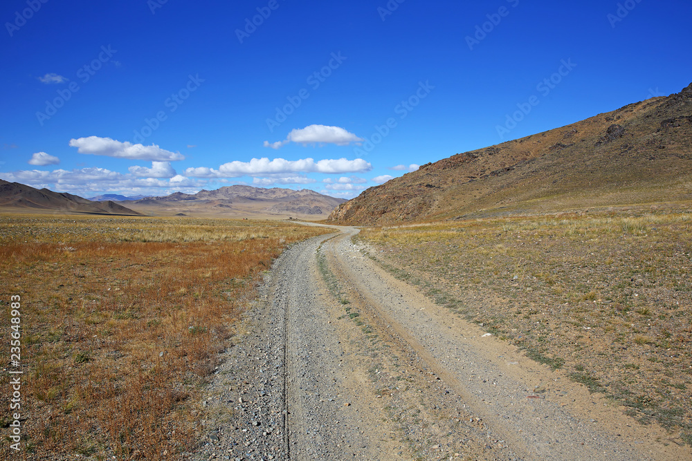 The rural road in the desert behind the mountain range with sky blue and cluods. Central Asia between the Russian Altai and Western Mongolia 