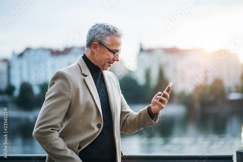 Mature businessman with smartphone standing by river in Prague city, taking selfie.