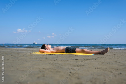 Fototapeta Naklejka Na Ścianę i Meble -  Teen  boy lies on yellow towel and sunbathes on the beach on the sea and sky background. Concept