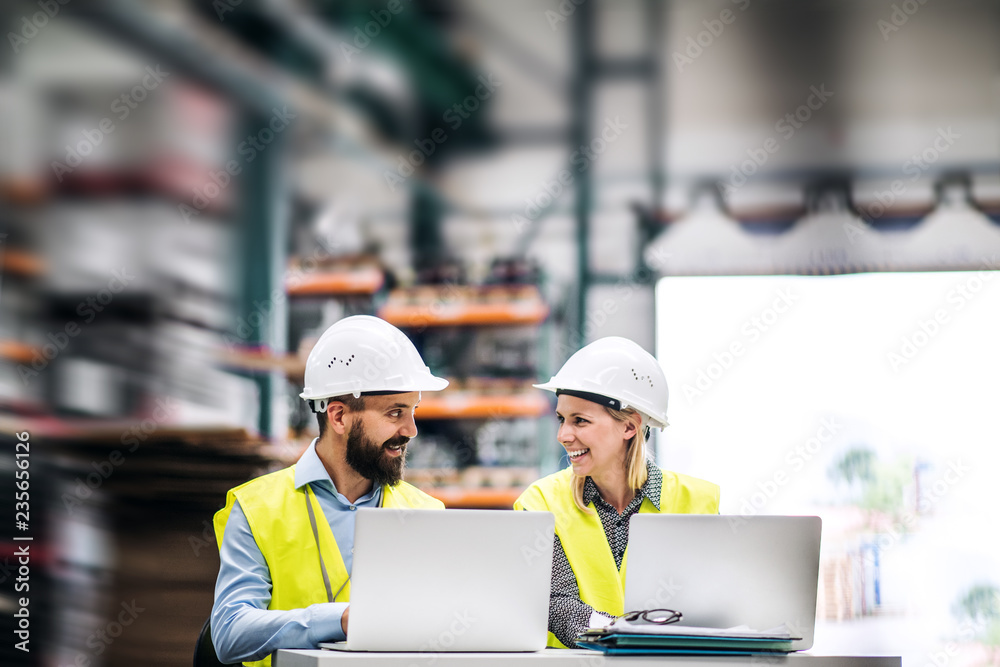 A portrait of an industrial man and woman engineer with laptop in a factory, working. A portrait of an industrial man and woman engineer with laptop in a factory, working.