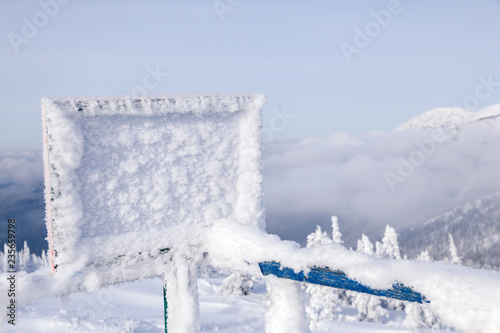 Rectangular wooden plate covered with hoarfrost on ski resort against the background of winter sunny scenic landscape  mountains  trees. Place for inscription  copyspace