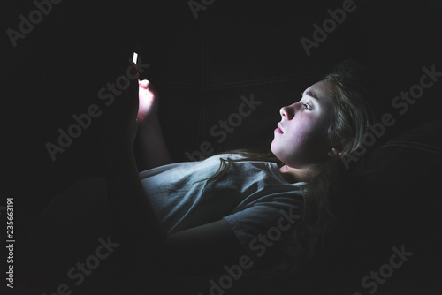 Depressed girl lying down on a couch in the dark while using her smartphone. The light from the screen is illuminating her face.