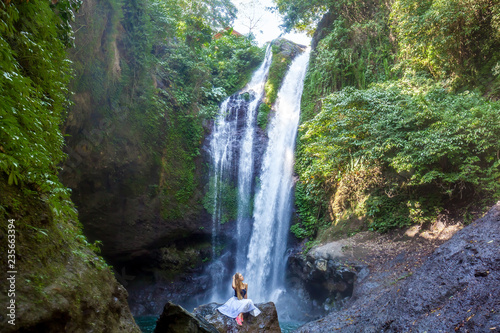 Young woman with long hair sitting on the rock in front of Aling-Aling waterfall among green tropical trees and plants on the north of Bali island, Indonesia