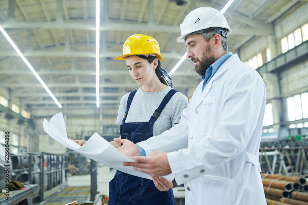 Side view portrait of female factory worker discussing work with mature engineer holding plans Side view portrait of female factory worker discussing work with mature engineer holding plans