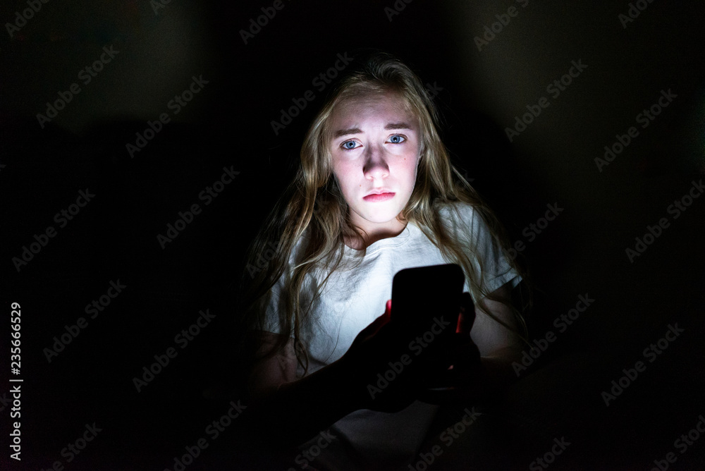 Upset girl sitting in the dark while using her smartphone. The light from the screen is illuminating her face. Upset girl sitting in the dark while using her smartphone. The light from the screen is illuminating her face.